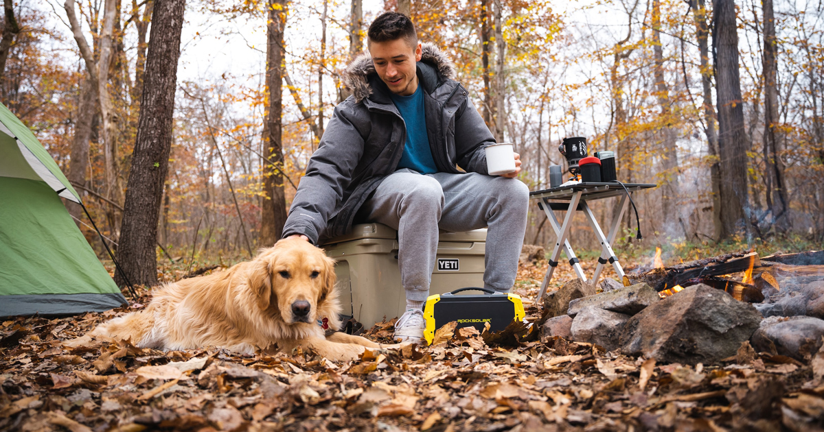 Man camping in forest sitting next to fire, petting golden retriever dog with portable power supply connected to electric appliances used for cooking.