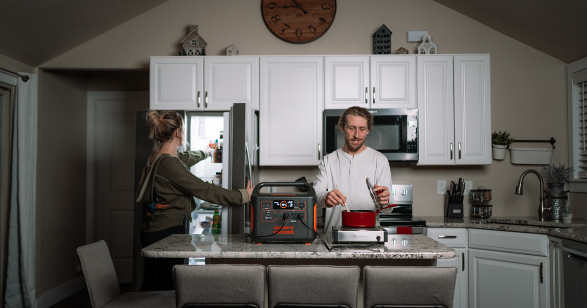 Dimly lit kitchen, woman reaching into refrigerator, man stirring food in a pot on an electric stove on counter plugged into portable battery.