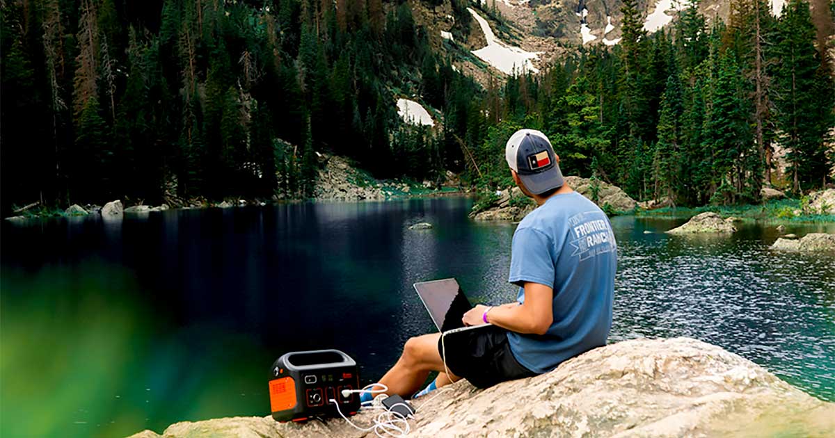 Man in mountains sitting on large rock overlooking lake while using laptop connected to portable power supply.